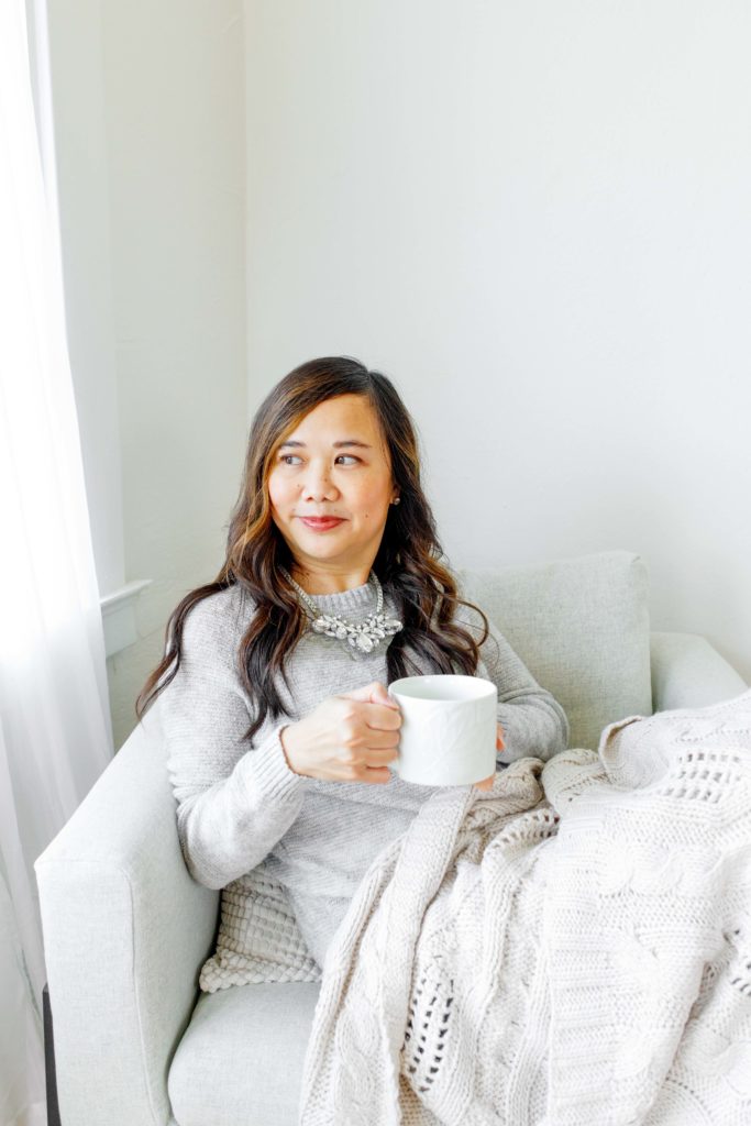 woman sitting in a grey chair with a blanket over her lap looking out the window and holding a mug of tea.