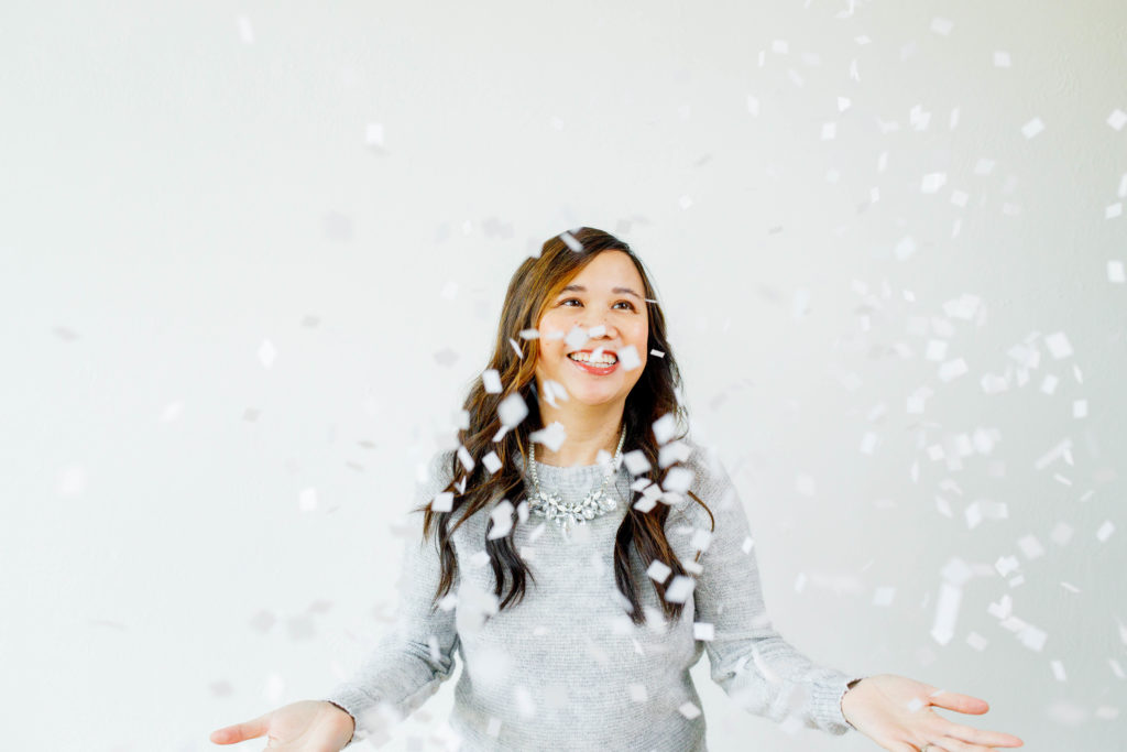 woman in a grey sweater and festive necklace smiling while throwing white squares of snow-like confetti in the air.