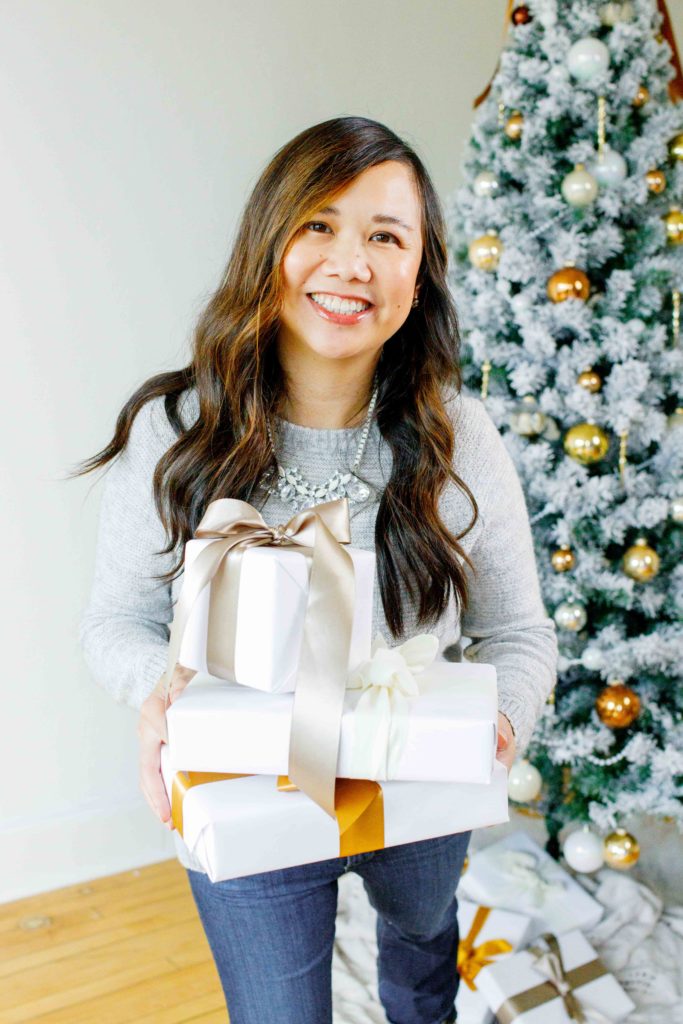 A woman holds white boxes with ribbon bows with a flocked Christmas tree in the background behind her.